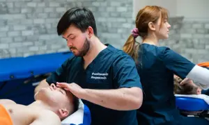 Fizjoterapeuta wykonuje masaż twarzy pacjentowi, w tle inna fizjoterapeutka.