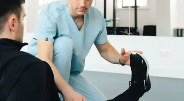 Fizjoterapeuta w jasnoniebieskim uniformie pomaga pacjentowi w ćwiczeniach rozciągających nogę.